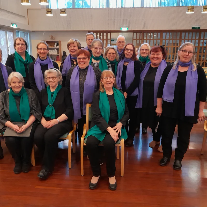 Suovanlahden kuoro ryhmäkuvassa, jäsenet pukeutuneet mustaan ja vihreisiin tai violetteihin huiveihin